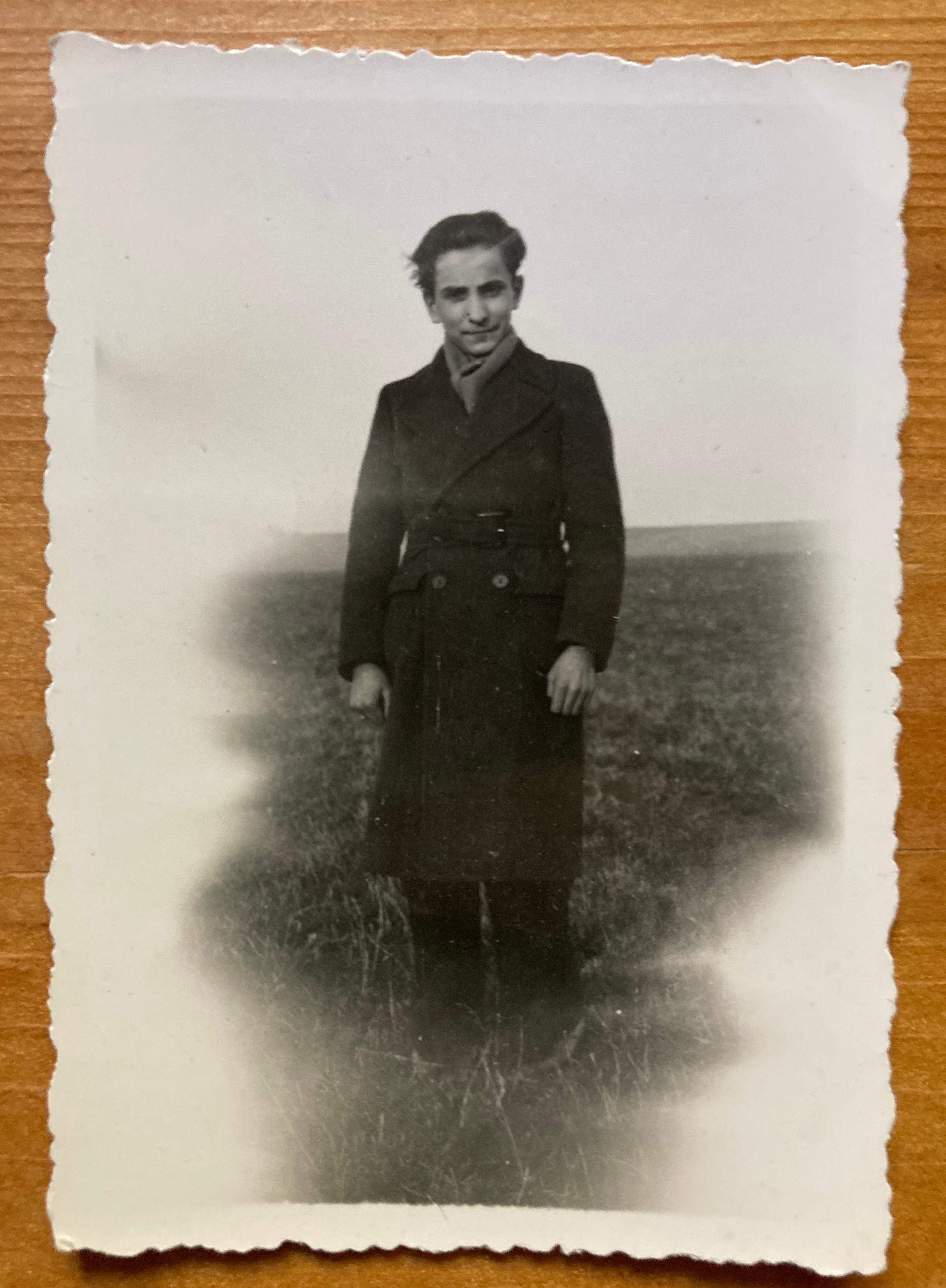 En svartvit fotografering av en ung man klädd i en lång mörk kappa, som står på en grön äng med neutrala bakgrundsdetaljer.